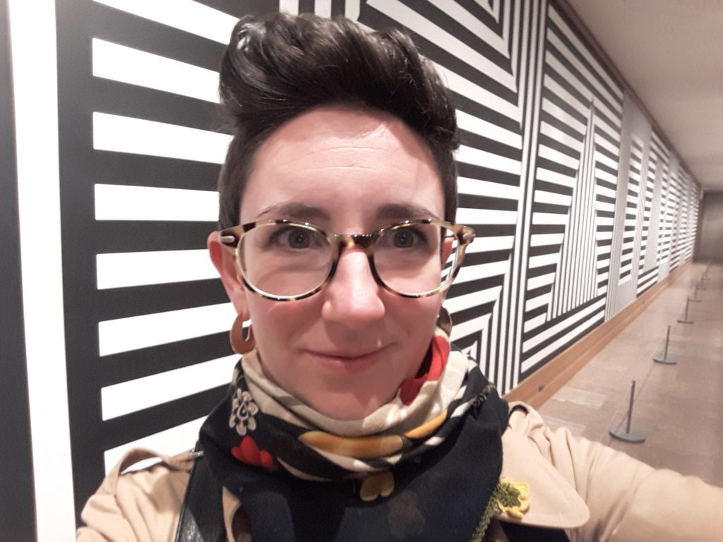 A 30 year old woman with short dark hair standing upright on her head stares straight into the camera as if she is taking a selfie. She wears tortise shell glasses, a colourful red, yellow and black scarf around her neck and is standing infront of a Sol Le Witt wall painting comprised black and white horizontal and then vertical stripes.