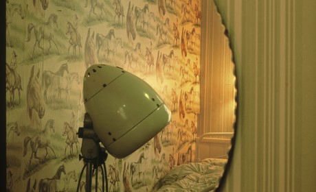 Melinda's Room by Leanora Olmi, 2015. An image of the reflection of a mirror showing the wall and part of a bed of a bedroom. The wallpaper is green and has a pattern of horses, and in front of it we see the head of a green old-fashioned lamp.